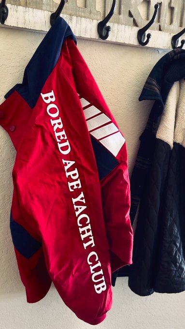 A red 'Bored Ape Yacht Club' jacket hangs on a wall hook rack alongside other outerwear.