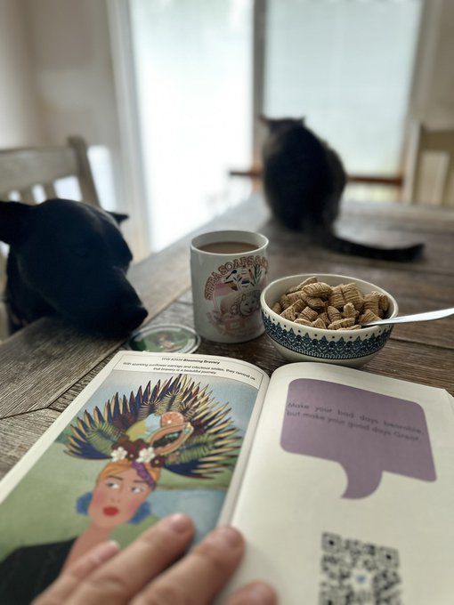 A cozy scene shows an open book on a wooden table alongside a coffee mug and a bowl of peanuts, with a black dog resting its head nearby and another cat sitting on the table.