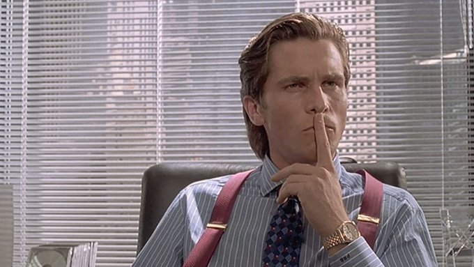 A man in a striped shirt, tie, and suspenders holds his finger to his lips in a contemplative, shushing gesture.