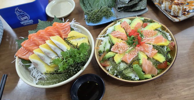 Two colorful sushi bowls filled with fresh salmon sashimi, avocado slices, egg, and crisp garnishes sit on a wooden table alongside a small dish of soy sauce.