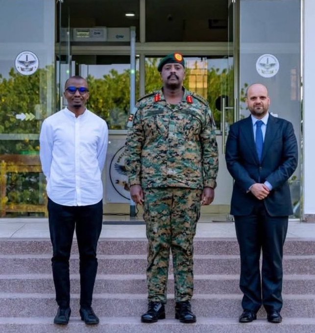 Three men pose for a photo on steps in front of a building entrance: one in a white shirt and sunglasses, a military officer in camouflage uniform with a green beret, and one in a dark business suit.
