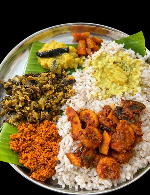 A traditional Sri Lankan rice and curry meal served on a metal plate lined with banana leaves, featuring white rice, a shrimp curry, a yellow dal, a green leafy dish, an orange coconut sambol, and other side dishes.