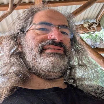 Close-up selfie of a man with long, wavy grey hair, a thick grey beard, and glasses, with a metal roof structure visible in the background.