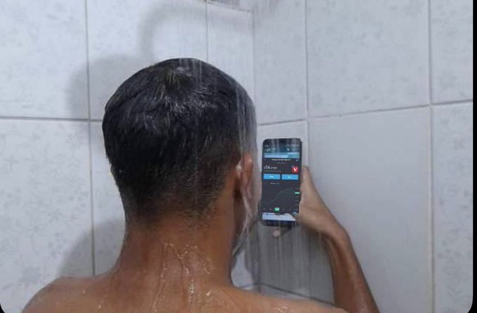 A man with wet hair takes a mirror selfie in the shower while checking a stock trading app on his phone, humorously illustrating the contradiction between knowing not to obsessively check investment portfolios and doing it anyway—even in the shower. The phone screen shows a stock chart with what appears to be a downward trend, emphasizing the anxious compulsion to monitor gains and losses.