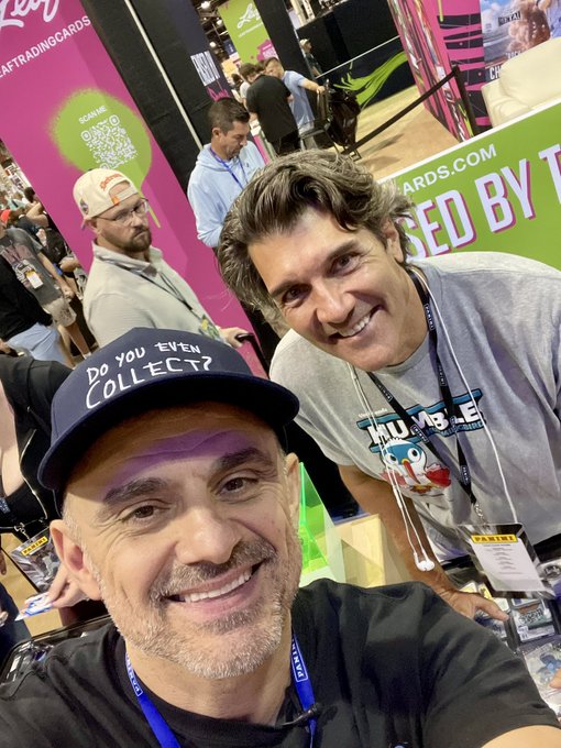 Two men posing for a selfie at what appears to be a trading card convention, with the foreground man wearing a "DO YOU EVEN COLLECT?" baseball cap and the background man wearing a "Double Dare" shirt.