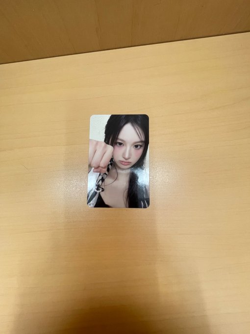 A photocard of a young woman with long dark hair posing with her fist near her face, resting on a light wood-grain surface.