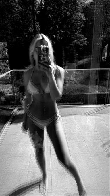 A black and white mirror selfie of a blonde woman in a bikini, standing on a tiled patio with trees visible in the background, her face obscured by her phone. The post suggests a casual midweek check-in, with the relaxed, summery photo complementing the low-key, introspective tone.