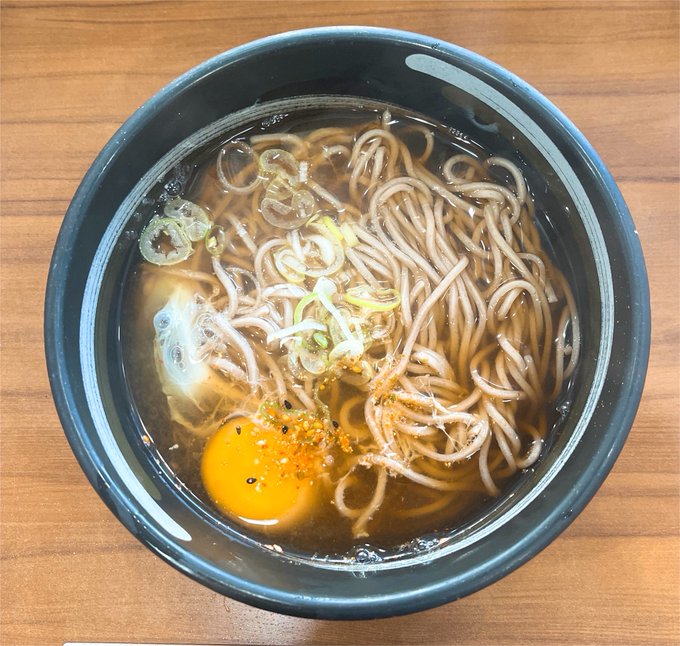 A bowl of Japanese soba noodles in clear broth, garnished with green onions and a raw egg yolk, served on a wooden table.