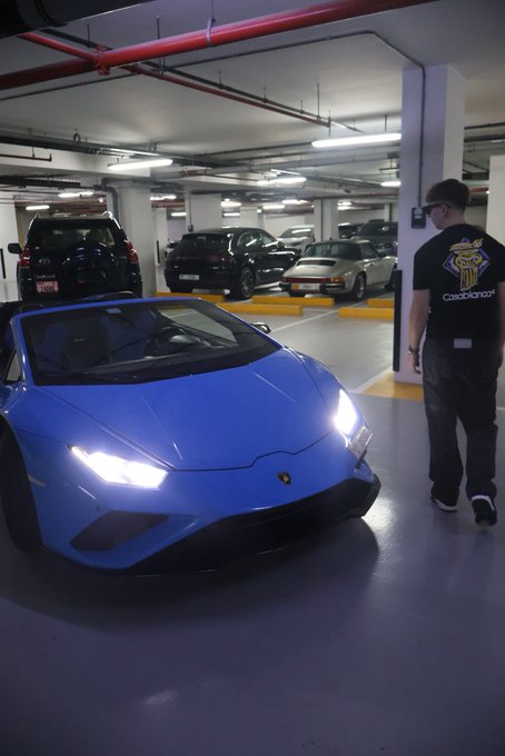 A blue Lamborghini Huracán is parked in an underground parking garage with its headlights on, while a man wearing a black "Catchshark" t-shirt walks past it, possibly indicating the car belongs to or is associated with the "GN" (Galpin) dealership.