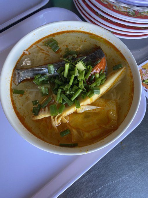 A bowl of Vietnamese fish soup topped with fresh green onions and herbs, served on a white tray next to a stack of decorative plates. The image complements the post, as the writer is enjoying a well-deserved meal after helping his wife with the grand opening of her new shop, wishing his followers a successful new week.