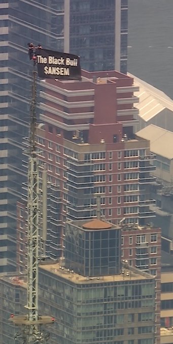 Workers atop the Empire State Building in NYC hoist a large digital billboard displaying "The Black Bull" alongside the ticker "$ANSEM" against the city skyline.
