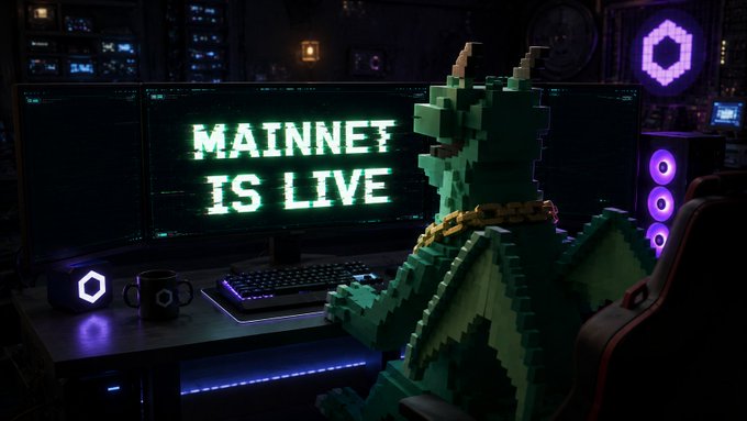 The image features a green dragon sitting at a computer desk, with the words "MAINNET IS LIVE" displayed above its head. The dragon appears to be playing video games or browsing the internet, as there are two monitors present on the desk. A keyboard and mouse can also be seen in front of the dragon's chair.