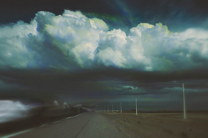 A dramatic landscape showing a dark road leading toward a massive, bright cloud formation beneath a stormy sky.