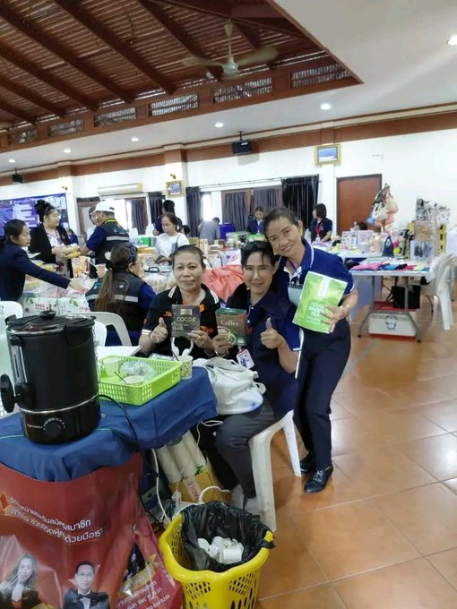 Three women in blue uniforms smiling and giving thumbs up while holding product packages, seated at a table with coffee dispensers and promotional materials in what appears to be a community event hall.