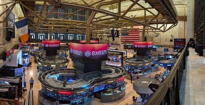 An elevated view of a trading floor shows multiple circular trading stations and large screens displaying the "exein" logo beneath an American flag.