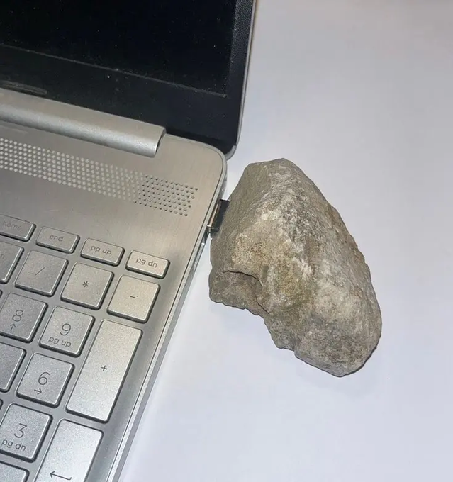 A gray rock propped up against the side of an open silver laptop on a white surface.