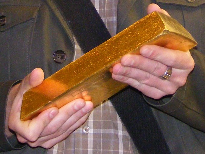 A man in a suit jacket and tie holds a very large gold bar between both hands, wearing a ring on his finger.
