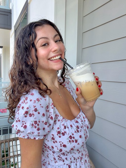 A smiling young woman with curly brown hair in a floral dress drinking an iced coffee through a straw on a porch.