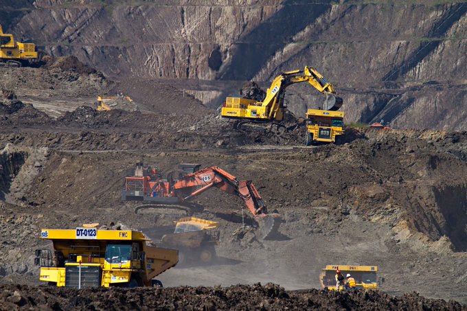 An open-pit mine scene with heavy machinery including yellow Komatsu excavators and bright orange Hitachi excavators working on terraced rock walls.
