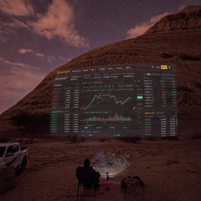 A surreal composite image showing a person in a hooded jacket sitting in a chair in a desert canyon at night, facing a massive transparent Binance trading interface for BTC/USDT that floats in the air, while a person in a pickup truck observes from the side.
