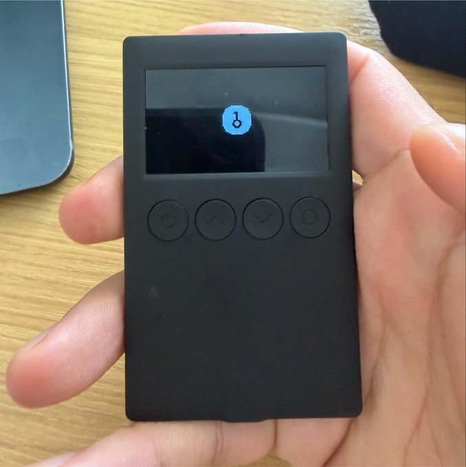 A hand holds a small black electronic device with four buttons (power, up, down, and another) below a small screen displaying a blue icon.