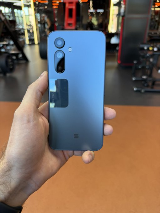 A hand holds up a blue smartphone showing its back camera module and a sticker reading 'HD TWS', appearing to be taken in a gym setting.