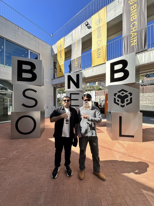 Two men wearing face masks and sunglasses pose in front of large cube-shaped letters spelling 'BSO' and 'BNB', with banners reading 'BNB CHAIN' hanging above them.