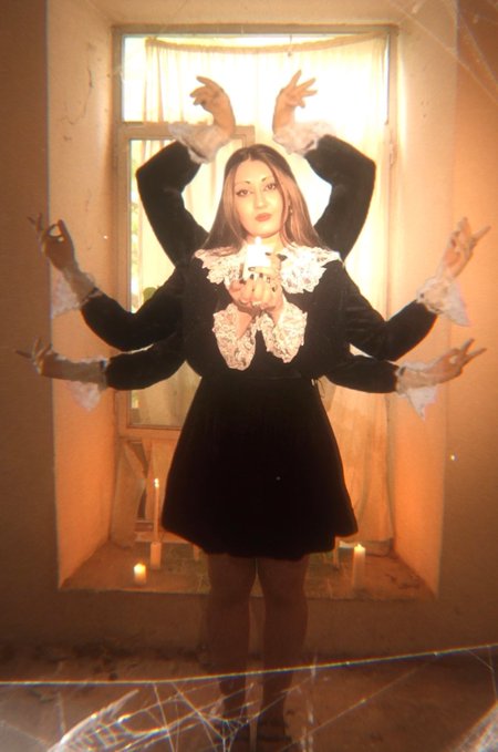 A woman with multiple arms appearing in a spider-like pose, wearing a black velvet dress with white lace collar, holding a lit candle, with cobwebs visible in the foreground, creating a mystical, Halloween-esque aesthetic.