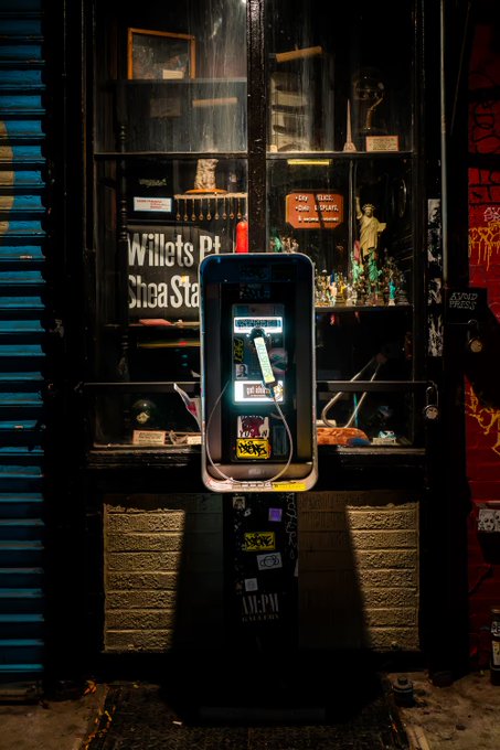 The image features a payphone booth with a sticker on its door, which reads "Collect calls iykyk". This means that the payphone allows users to make international phone calls for a fee. The payphone is located outside of a store or a building and has a window display showcasing various items inside.
