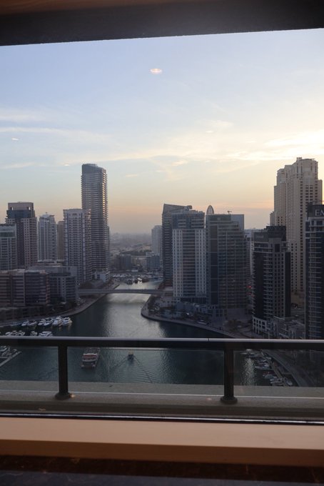 A waterfront city view at dusk, taken from a balcony, with high-rise buildings lining both sides of a canal where boats are docked.

The image shows the same cityscape but looking down on it from above, with the text "GM" visible on one of the buildings—a perfect visual response to anyone saying "good morning" back, since this view greets the viewer from a high vantage point.