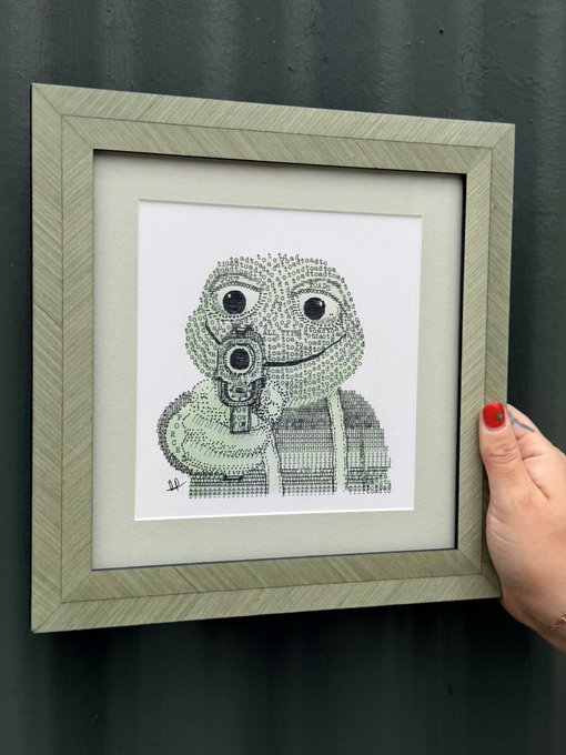 A framed piece of text-art depicting a frog holding a gun, created entirely from tiny characters, with a hand featuring red nail polish holding the frame.