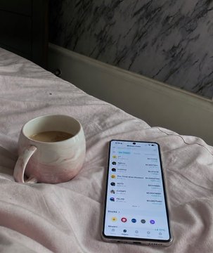 A morning scene featuring a ceramic coffee mug alongside a list of Zealy earning opportunities displayed on a smartphone resting on white bedding, complementing the post's message about accessible Web3 income streams like the Injective Zealy Program for small creators.