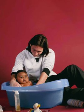 A woman in a white shirt bathes a baby in a blue tub against a red background, with baby supplies visible.