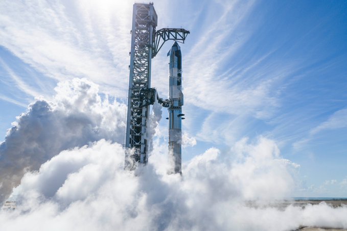A SpaceX Starship rocket stands on a launch pad with its booster, surrounded by billowing white smoke and vapor against a blue sky with wispy clouds.