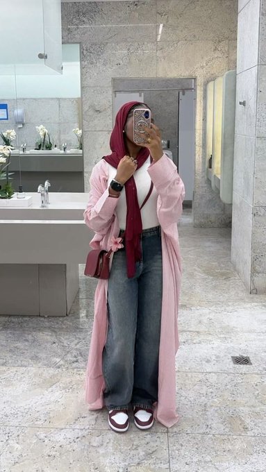 A woman wearing a pink open robe over jeans and a maroon hijab takes a mirror selfie with her phone in a marble-tiled restroom.