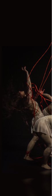 An artistic photograph of a dancer in a flowing dress entangled in red ribbons against a dark background, reaching upward dramatically.