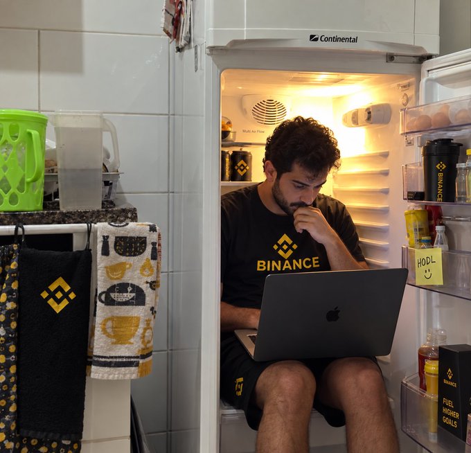A young man wearing a Binance-branded shirt sits inside an open refrigerator, focused on his Apple laptop in a creative makeshift workspace. Visible text includes "BINANCE" on his shirt, a "Continental" logo on the fridge, and a sticky note reading "HODL" with a smiley face—ironically illustrating the caption about how even a fridge has become a suitable place to work, likely tying into themes of crypto trading and the "HODL" mentality.