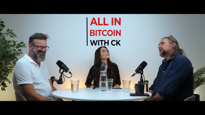 The image features three people sitting around a table, with two men wearing glasses and one woman. They are engaged in a conversation about Bitcoin, as indicated by the text "All In Bitcoin With CK". The scene takes place in an office setting, which is suggested by the presence of potted plants on either side of the room.