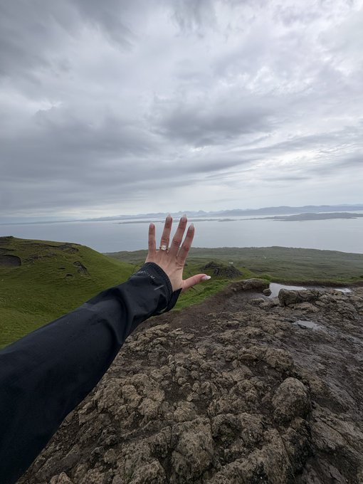 A hand reaches out over a dramatic, cloud-covered coastal landscape, displaying an engagement ring to celebrate the "She said yes!" moment.