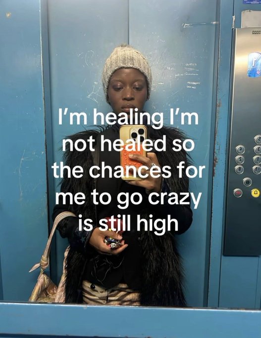 A woman wearing a beige beanie and a black furry vest takes a mirror selfie in an elevator with blue walls, while text overlaid reads: "I'm healing I'm not healed so the chances for me to go crazy is still high."