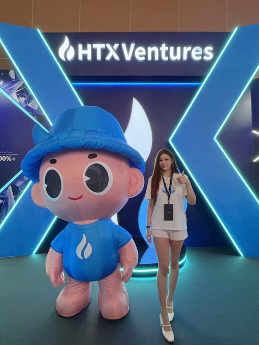 A woman in a white shirt and shorts stands next to a pink and blue mascot wearing an 'HTX' shirt, in front of an illuminated 'HTXVentures' display.