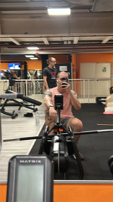 A muscular, tattooed man in a pink shirt takes a mirror selfie with his phone while sitting on a rowing machine at a gym, with another man standing in the background.