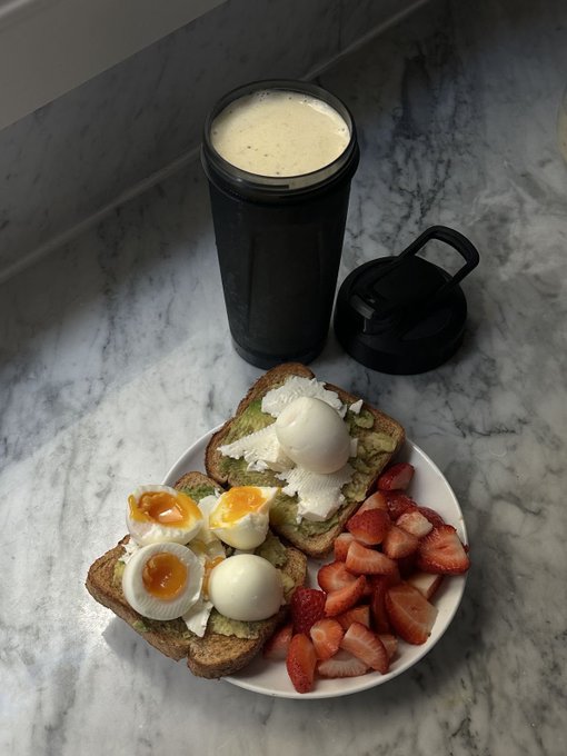 A breakfast plate on a marble counter featuring avocado toast topped with soft-boiled eggs and feta cheese, accompanied by sliced strawberries and a black tumbler with creamy beverage.