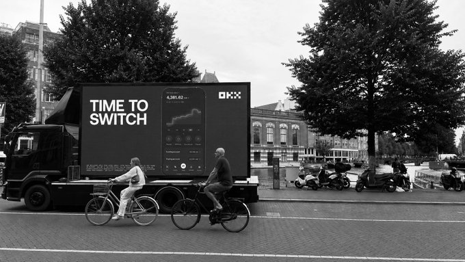 A black-and-white street scene shows two cyclists passing a mobile billboard truck displaying an OKX advertisement featuring a smartphone mockup, the text "TIME TO SWITCH," and a price chart reading "4,381.62," promoting OKX's MiCA compliance milestone referenced in the caption.