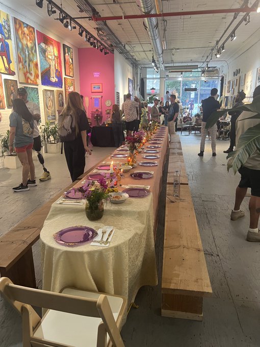 An indoor gallery space with colorful artwork on the walls, where people are gathered around a long communal table decorated with flowers and purple plates.