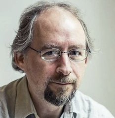 A middle-aged man with graying hair, a goatee, and wire-rimmed glasses looks directly at the camera, wearing a light beige collared shirt against a soft, neutral background, appearing as a thoughtful portrait-style photo of cypherpunk legend Adam Back, the Bitcoin advocate referenced in the accompanying post about his "smash buying" of Bitcoin during a current buying opportunity.