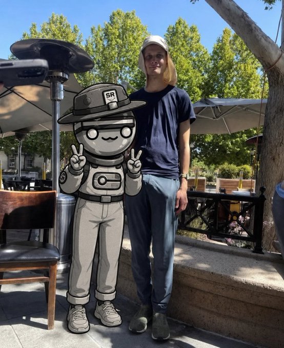 A man wearing a white bucket hat, black t-shirt, and gray pants stands outdoors under umbrellas next to a life-size cardboard cutout of an anime-style character in a gray military outfit with "SR" on its cap, making peace signs.