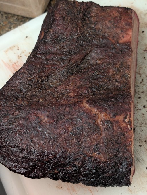 A large piece of smoked brisket with a dark, crusty bark rests on a white cutting board.