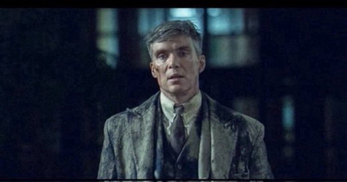 A cinematic shot of a man with tousled hair and dirt on his face, wearing a worn suit and tie, appearing distressed in a dimly lit setting.