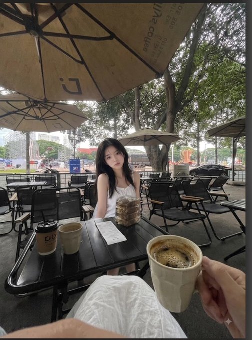 A first-person perspective photo at an outdoor café showing a young woman with long dark hair sitting across a table, with a coffee cup held in the foreground and pastries between them under a large umbrella.